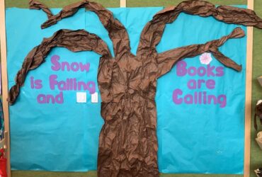 A display with a brown tree in front of a blue background. Around the tree are the letters in purple spelling "Snow is Falling and Books are Calling".