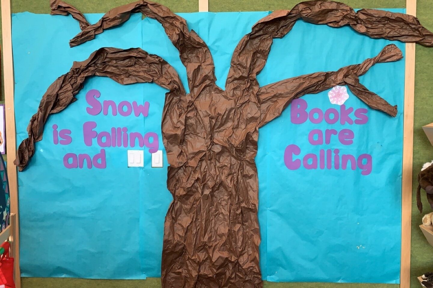 A display with a brown tree in front of a blue background. Around the tree are the letters in purple spelling "Snow is Falling and Books are Calling".