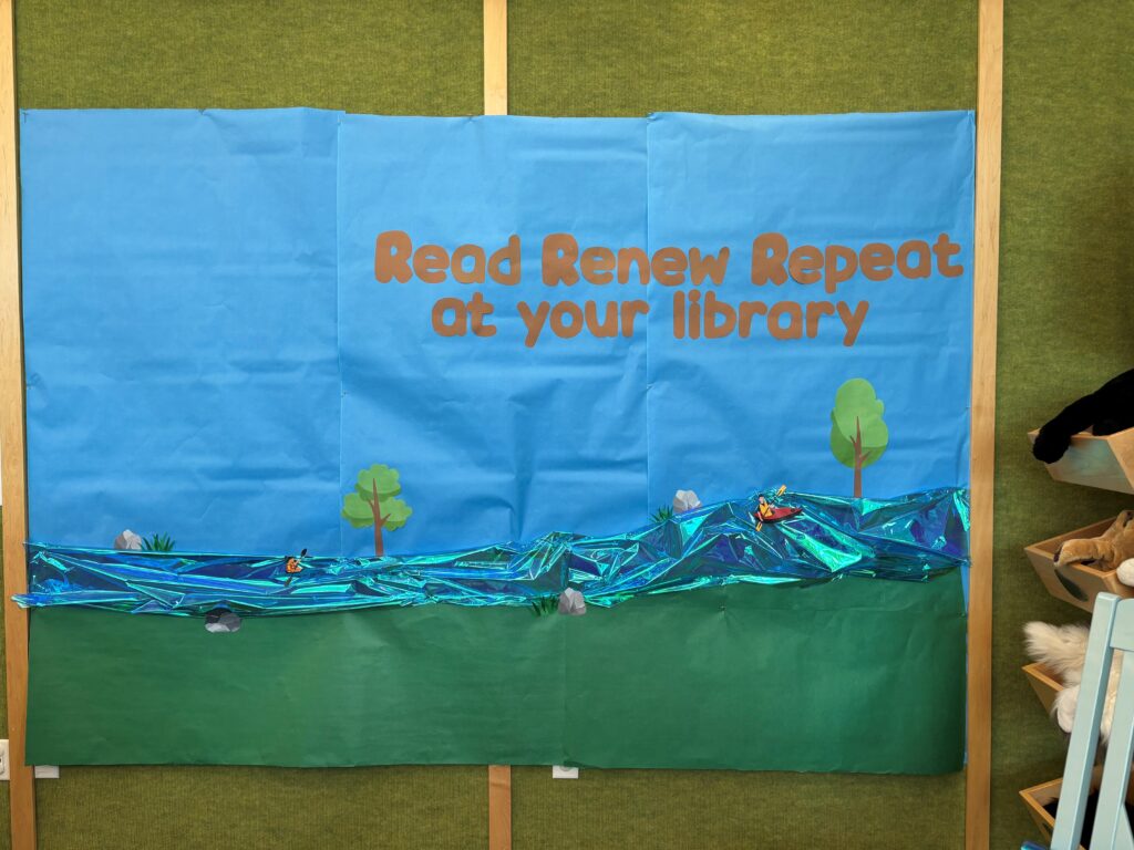 A display on the wall with blue paper on top and green on the bottom separated by a river. The words Read Renew Repeat at your library are above the river.