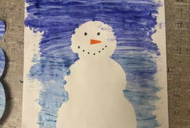 A snowman craft with blue paint behind it and is white as if made of snow.